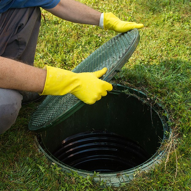 Man in overalls opening green septic tank lid with protective gloves.