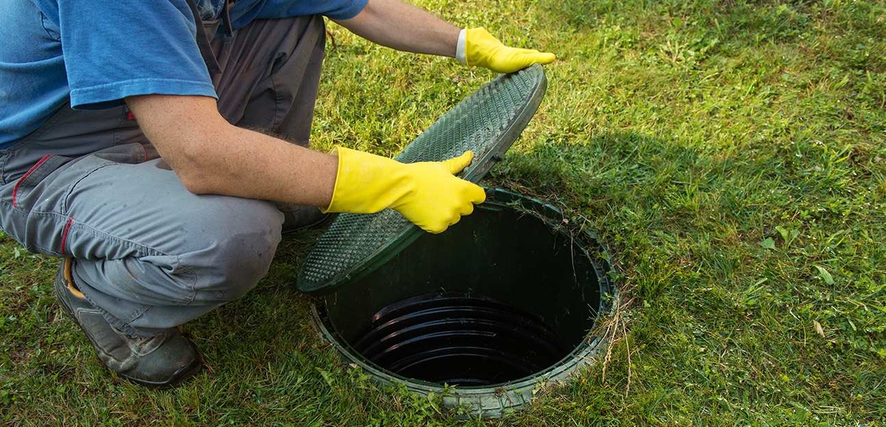 Man in overalls opening green septic tank lid with protective gloves.