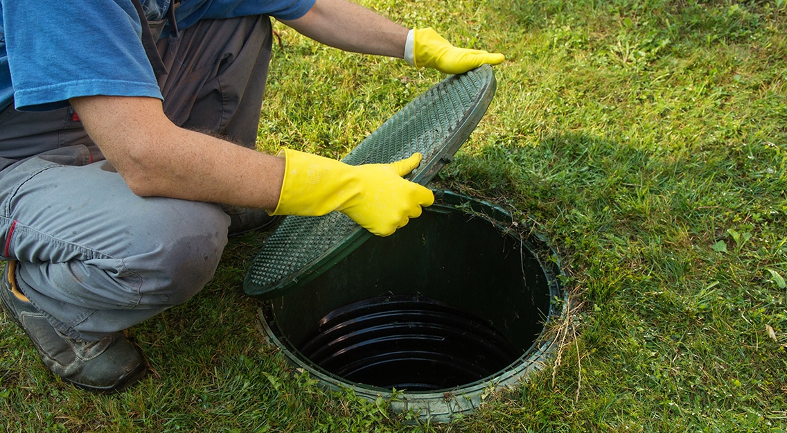 Man in overalls opening green septic tank lid with protective gloves.